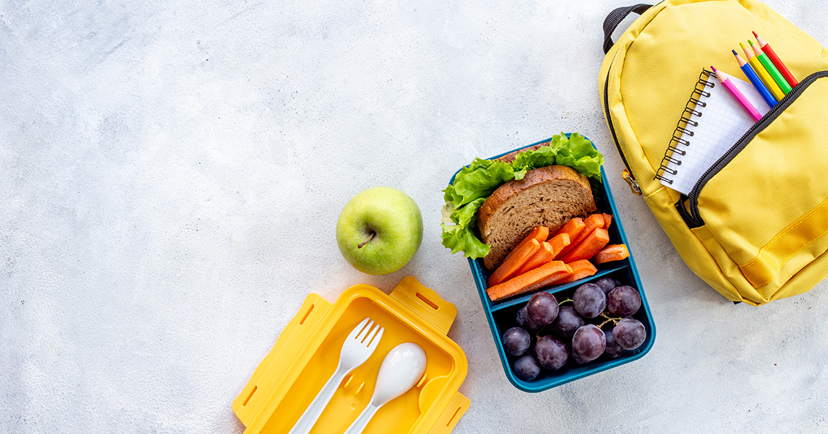 Lunchbox with a sandwich, carrots, and grapes beside a green apple and yellow backpack.